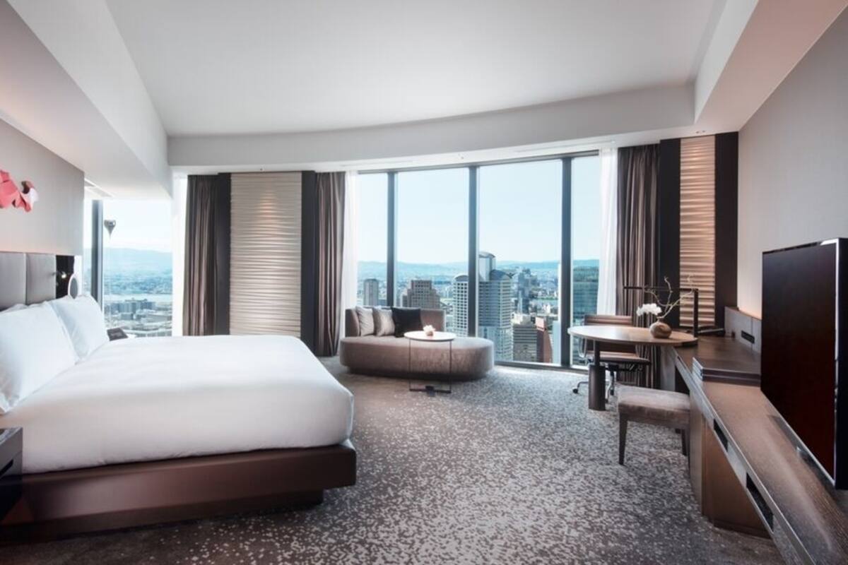 Ein modernes Hotelzimmer mit einem großen Bett, einem Schreibtisch, einem Sessel und einem Fernseher. Das Zimmer bietet Blick auf die Stadt.