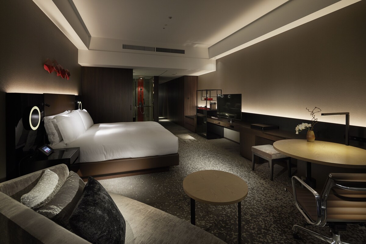 A modern hotel room with a large bed, a sofa, a small round table, a TV, and a desk with a computer.
