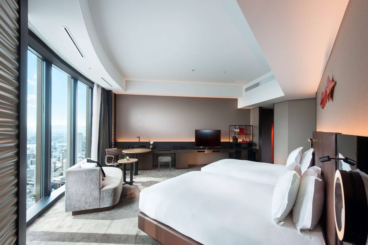 A modern hotel room with a large bed, a desk, a chair, a TV, and a view of the city.