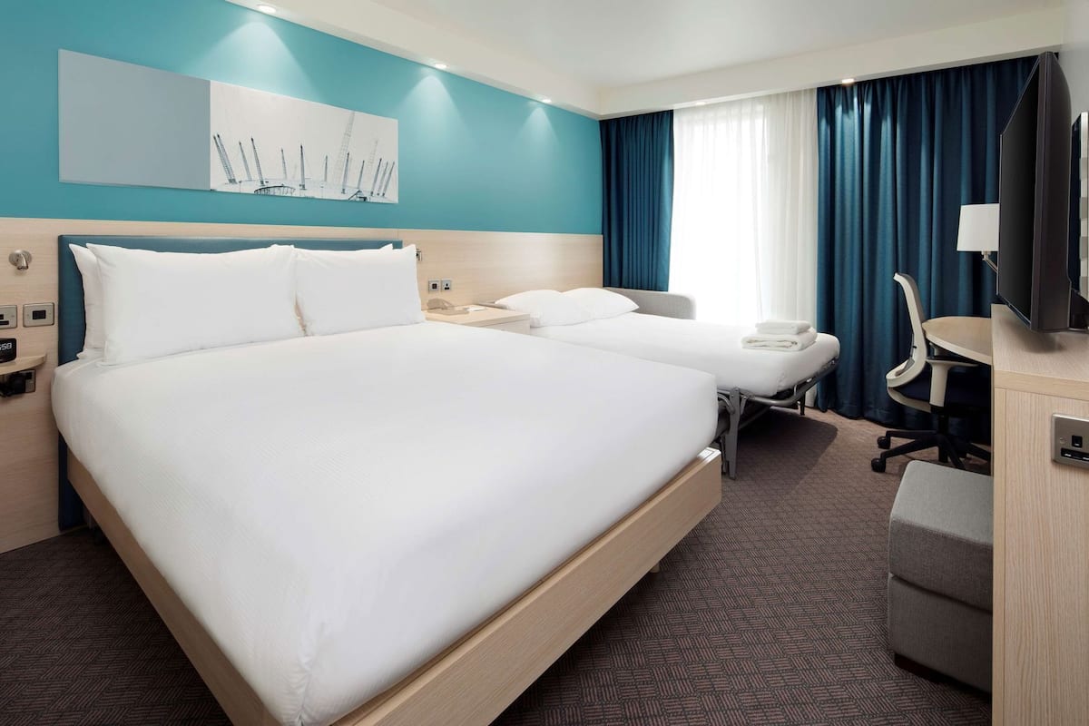 A hotel room with a large bed, a smaller bed, a desk with a chair, a TV, and a painting on the wall.