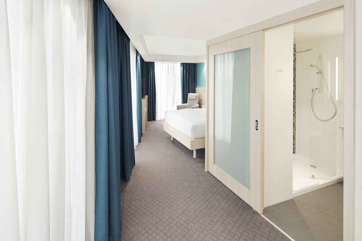 A modern hotel room with a bed, a seating area, and a glass-enclosed shower.