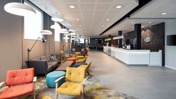 Una moderna hall dell'albergo con reception, area relax con sedute colorate e un'ampia finestra che lascia entrare luce naturale.
