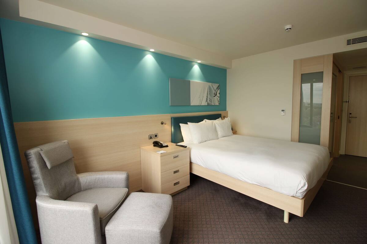 A hotel room with a bed, a chair, a nightstand, and a wall-mounted artwork.