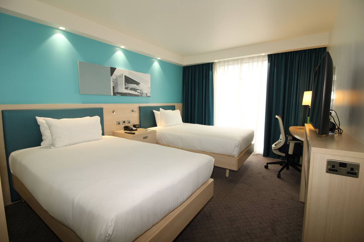 A hotel room with two single beds, a desk with a chair, a flat-screen TV, and a window with curtains.