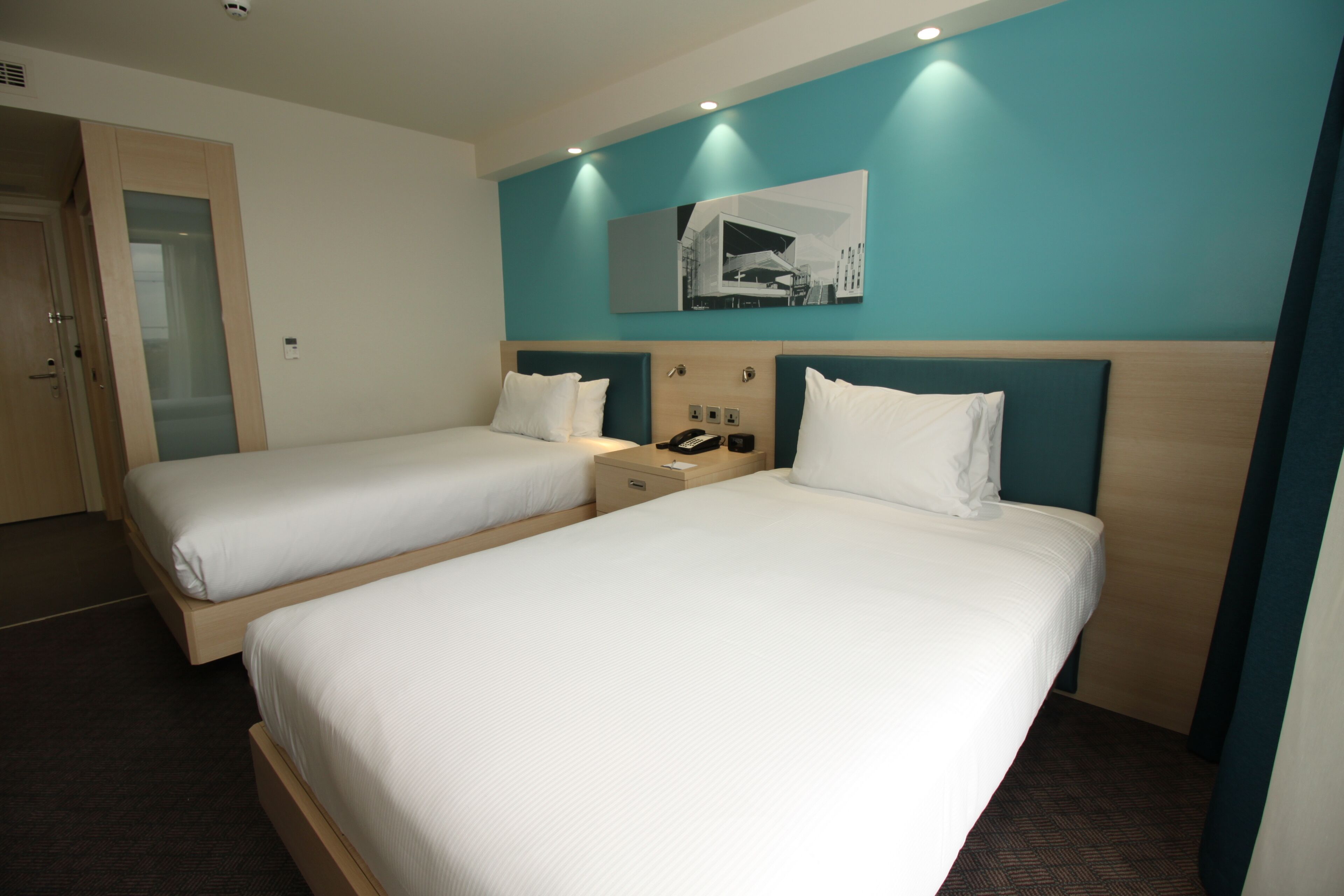 A hotel room with two beds, a nightstand with a phone, and a wall-mounted picture.
