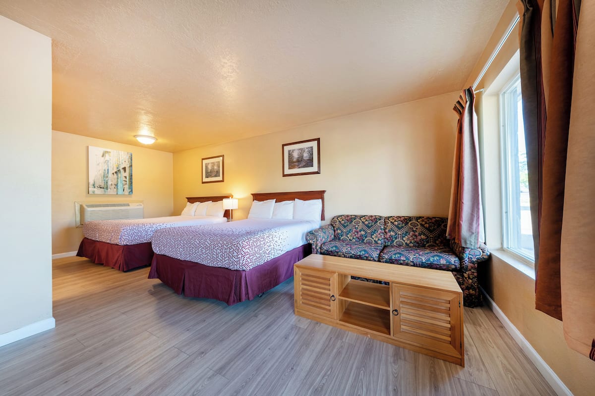 A hotel room with two beds, a sofa, a wooden side table, and a window with curtains.
