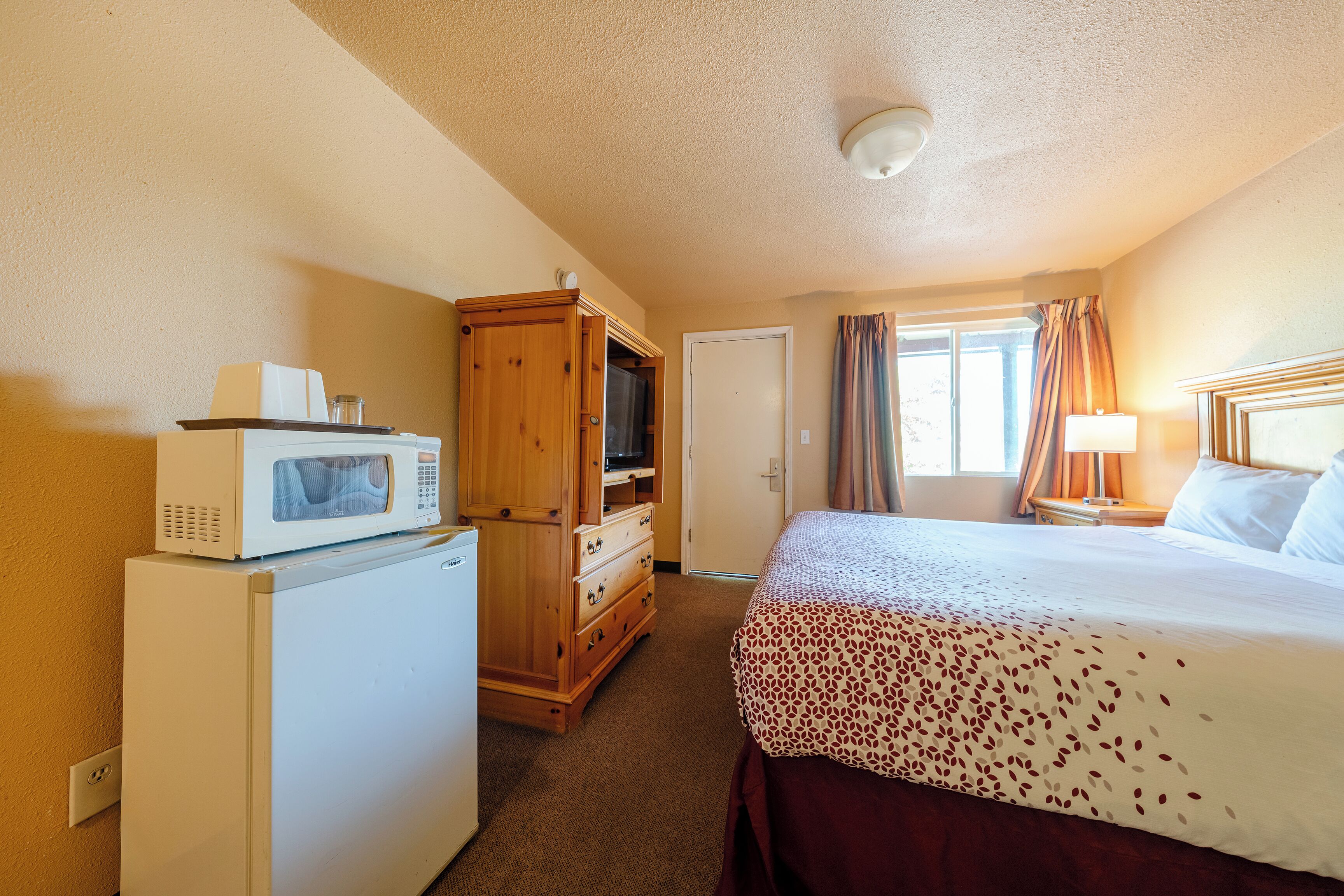 Une chambre d’hôtel avec un lit, un four à micro-ondes, une commode en bois, une porte et une fenêtre avec des rideaux.