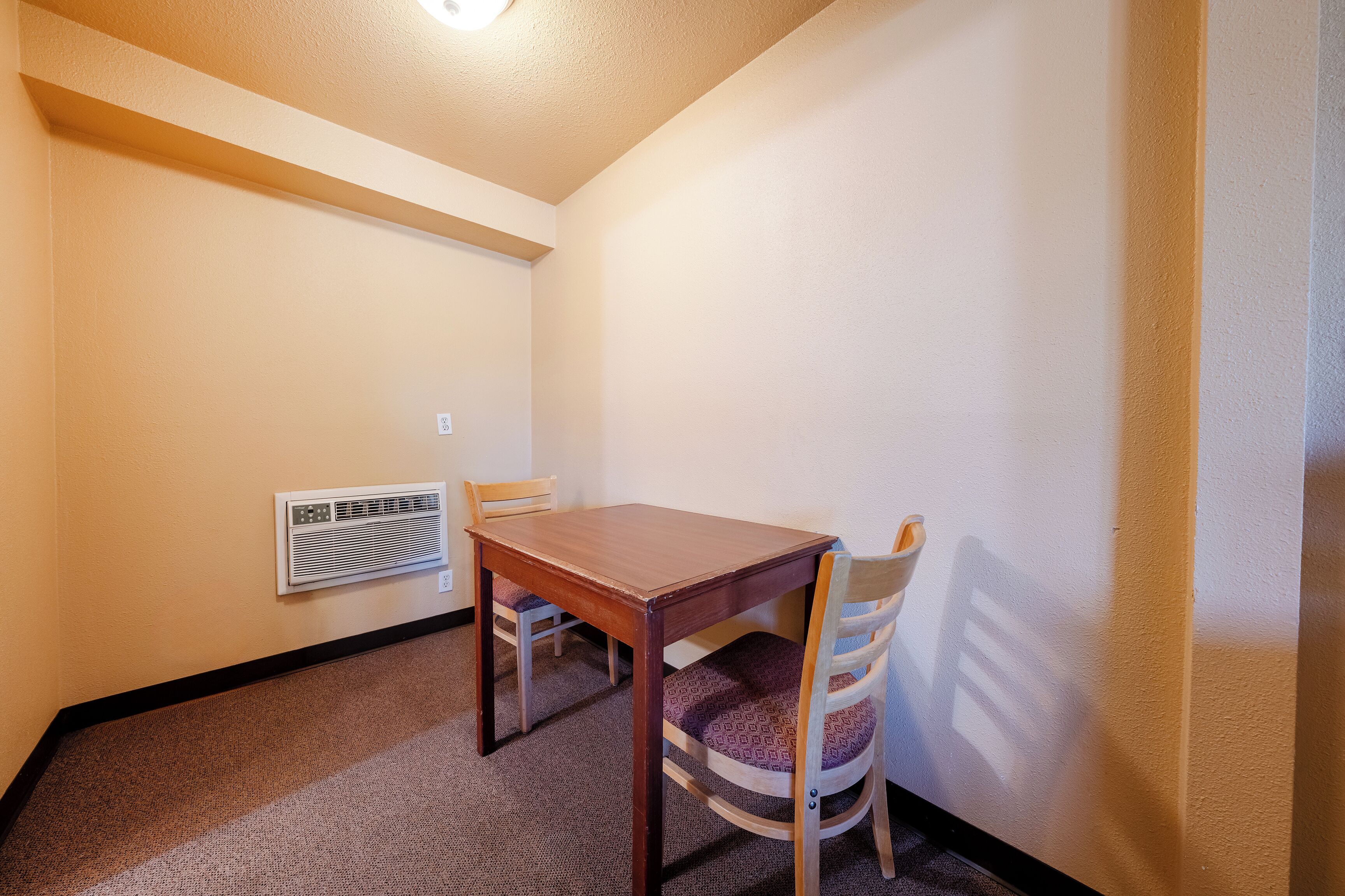 A small, carpeted room with a wooden table and two chairs, a wall-mounted heater, and a ceiling light.