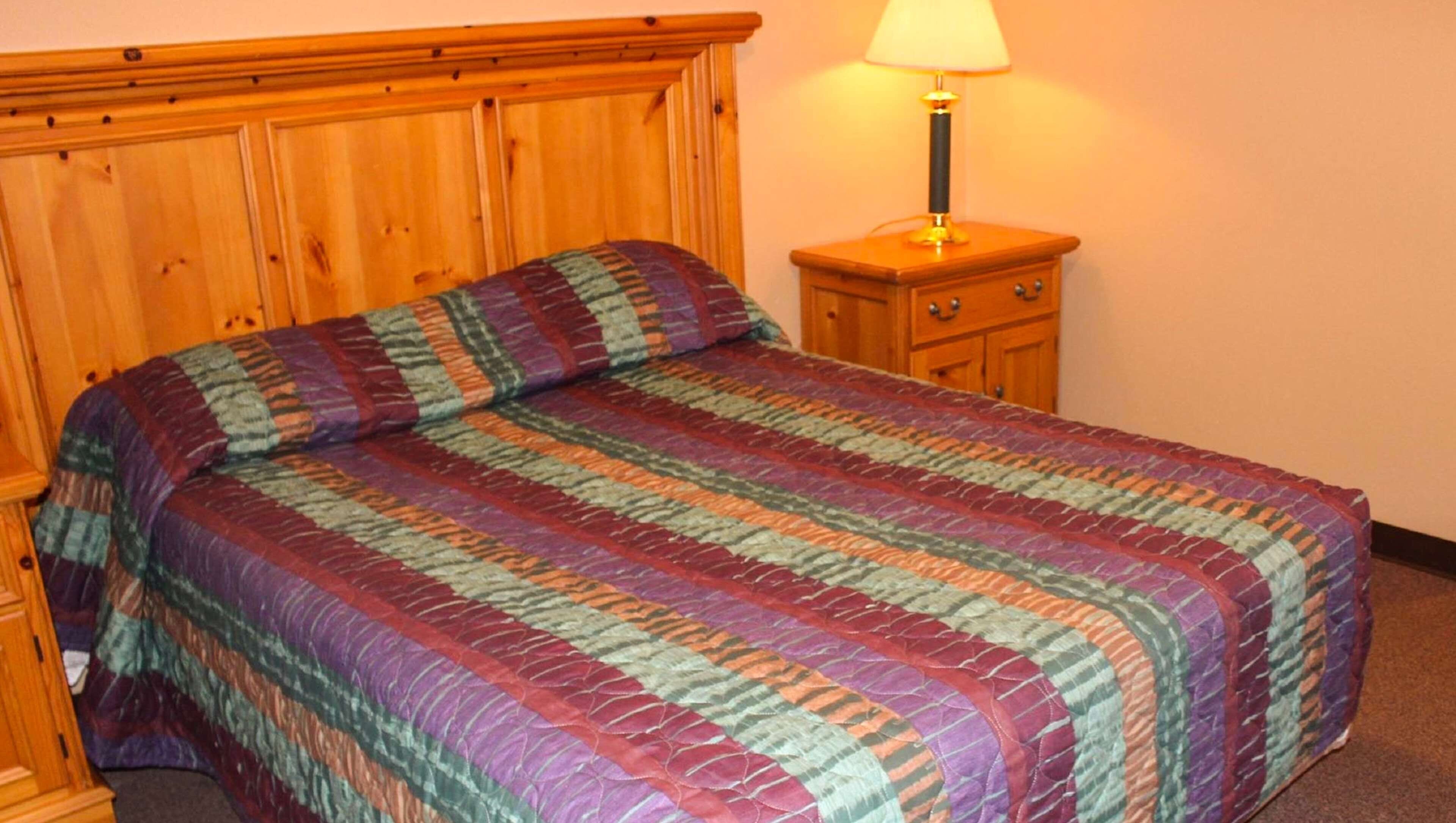 Una cama con un colcha de colores, un cabecero de madera, un mesita de noche con lámpara y cajones de madera.