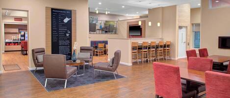 A spacious hotel lobby with a wooden floor, seating areas, a bar, and a television screen.