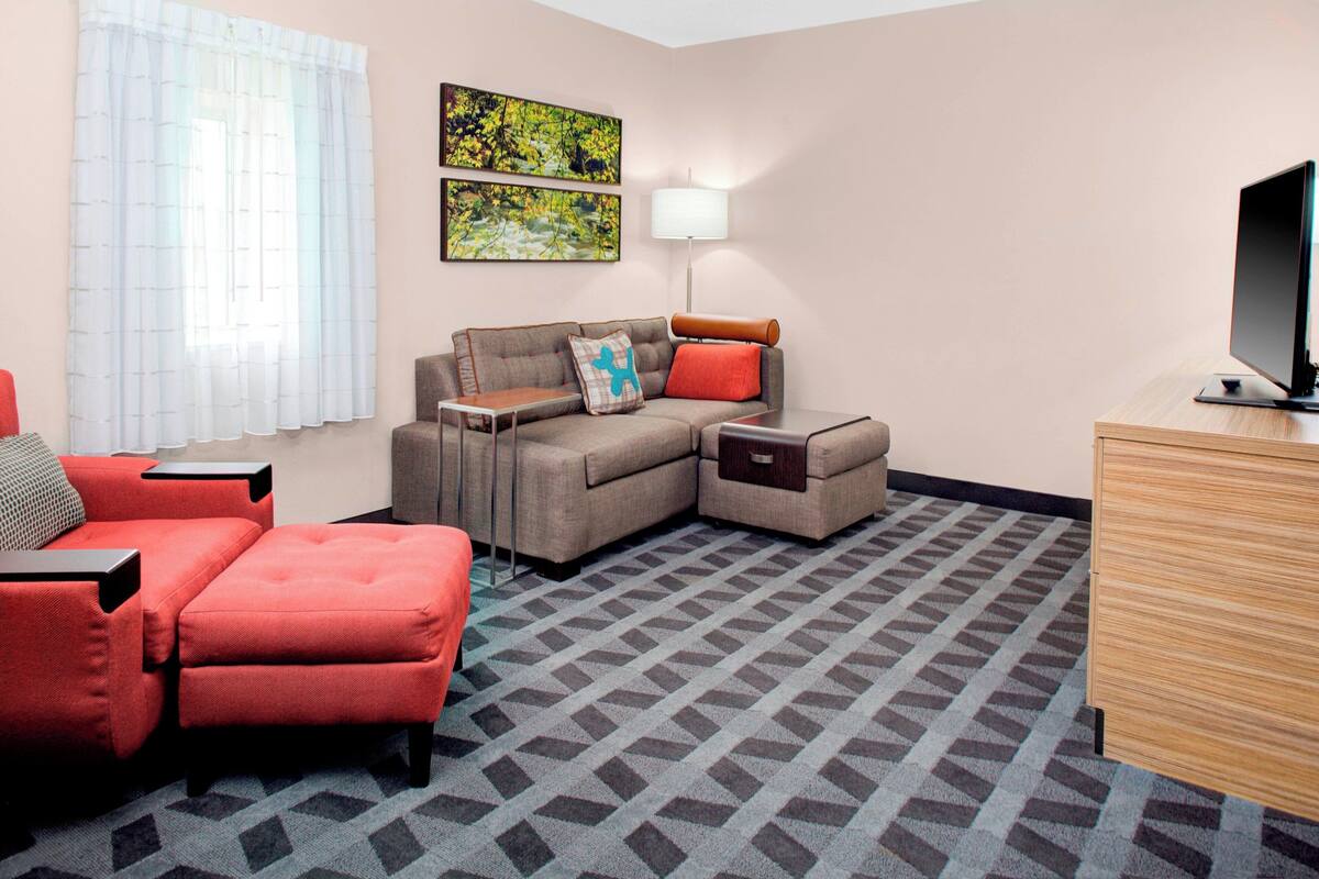 A hotel room with a sofa, a red armchair, a small table, a lamp, a TV, and a patterned carpet.