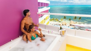 Um casal em uma banheira numa varanda com vista para o oceano e uma parede rosa.