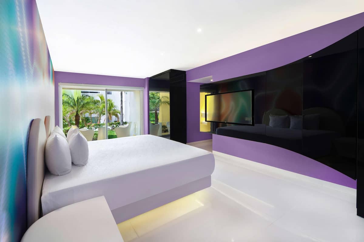 A modern bedroom with a large bed, a TV mounted on the wall, and a view of palm trees outside.