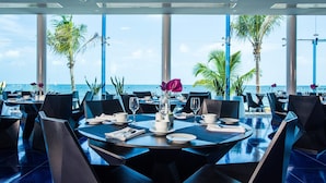 Um restaurante com mesas postas para refeições, grandes janelas e vista para o mar.