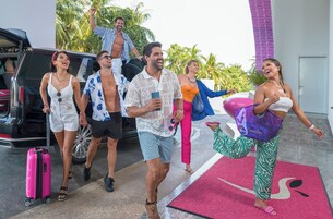 A group of people walking towards a building with a pink and white color scheme, palm trees in the background, and a car parked nearby.