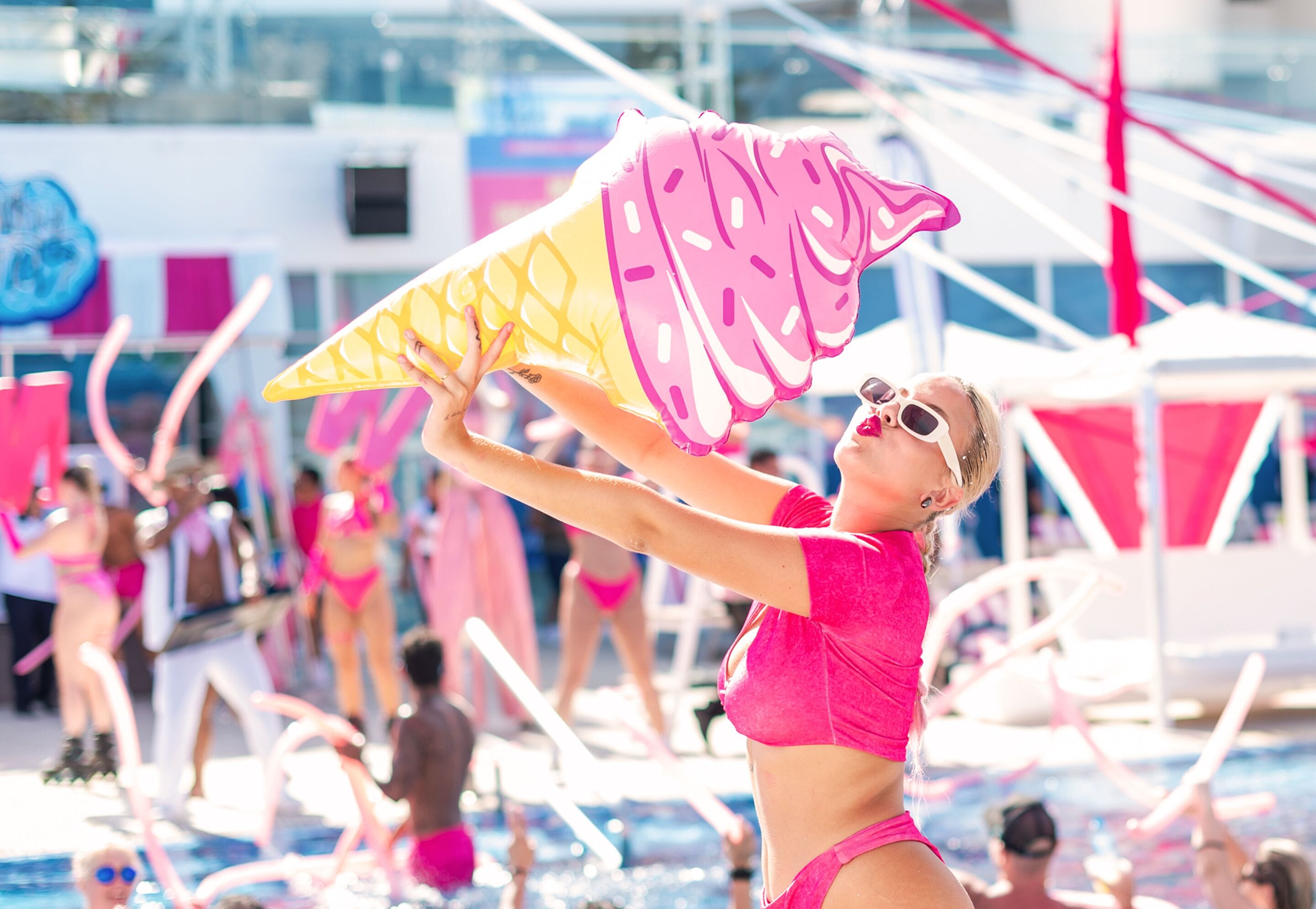 A person holding a large, colorful kite in a pool area with other people and a boat in the background.