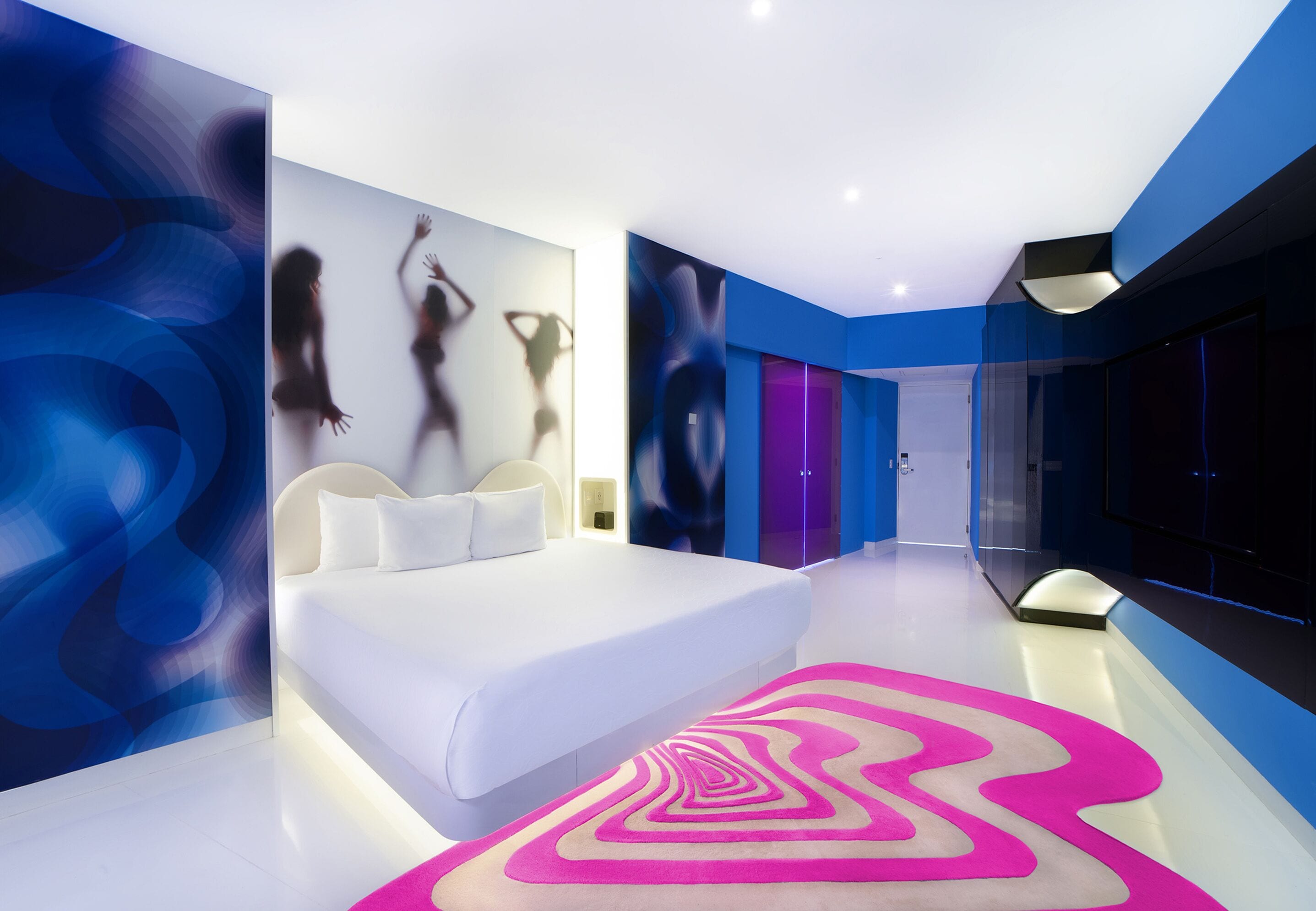 Une chambre d’hôtel moderne avec un grand lit, un tapis rose vif et des œuvres d’art abstraites accrochées au mur.