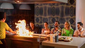 Un restaurante con un cocinero preparando un flambé, comensales sentados en una mesa con varios platos, y un papel tapiz decorativo al fondo.