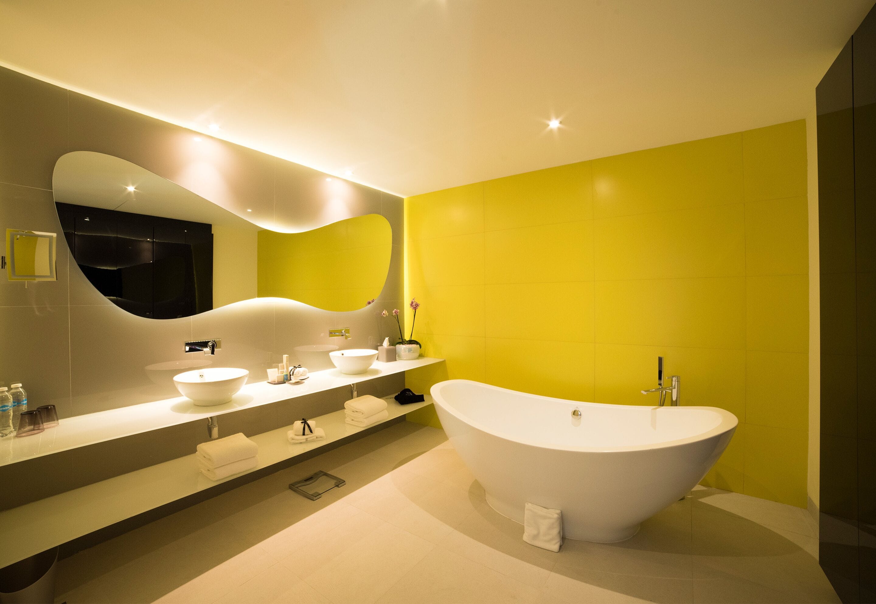 A modern bathroom with a freestanding bathtub, a large mirror, and a yellow accent wall.