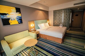 A hotel room with a bed, a sofa, a small table, a nightstand, a wall-mounted lamp, and a large abstract painting. - Holiday Inn Express Yingkou Onelong Plaza by IHG (Yingkou)