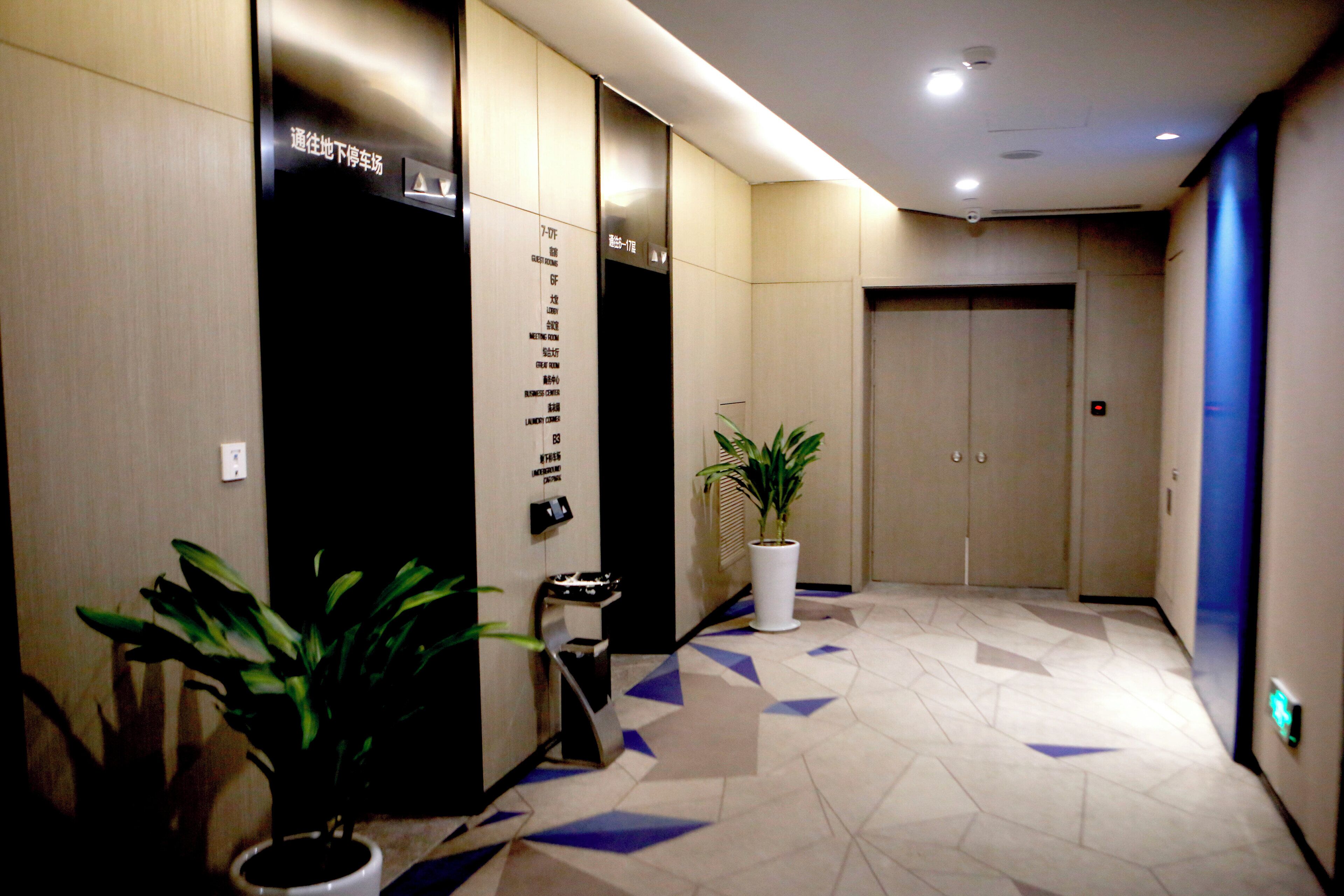 A hotel corridor with a geometric patterned floor, potted plants, and a reception area with a black elevator.