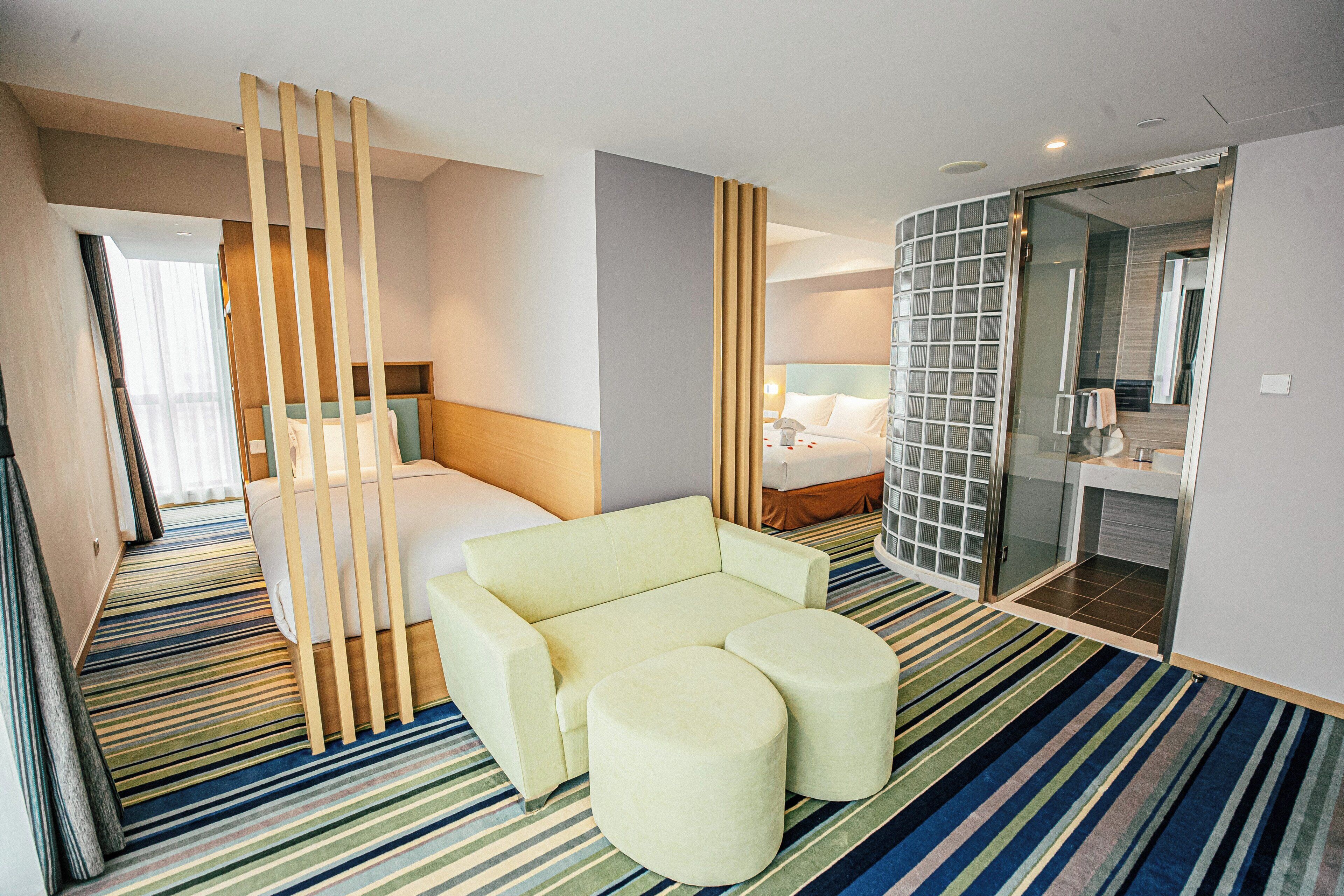 A hotel room with a bed, a sofa, a small table, and a bathroom with a glass partition.
