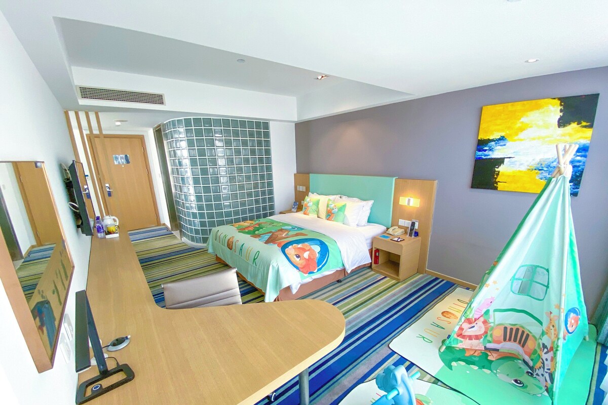 A hotel room with a bed, a desk, a chair, a mirror, a window, and a painting on the wall.