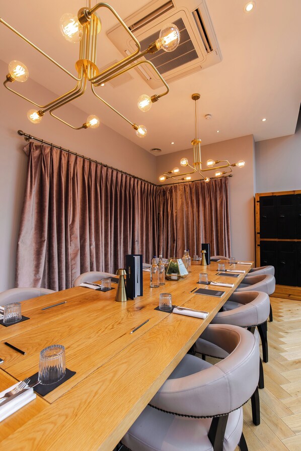 Un salón de conferencias con una larga mesa de madera, sillas tapizadas en gris y una lámpara de techo moderna.