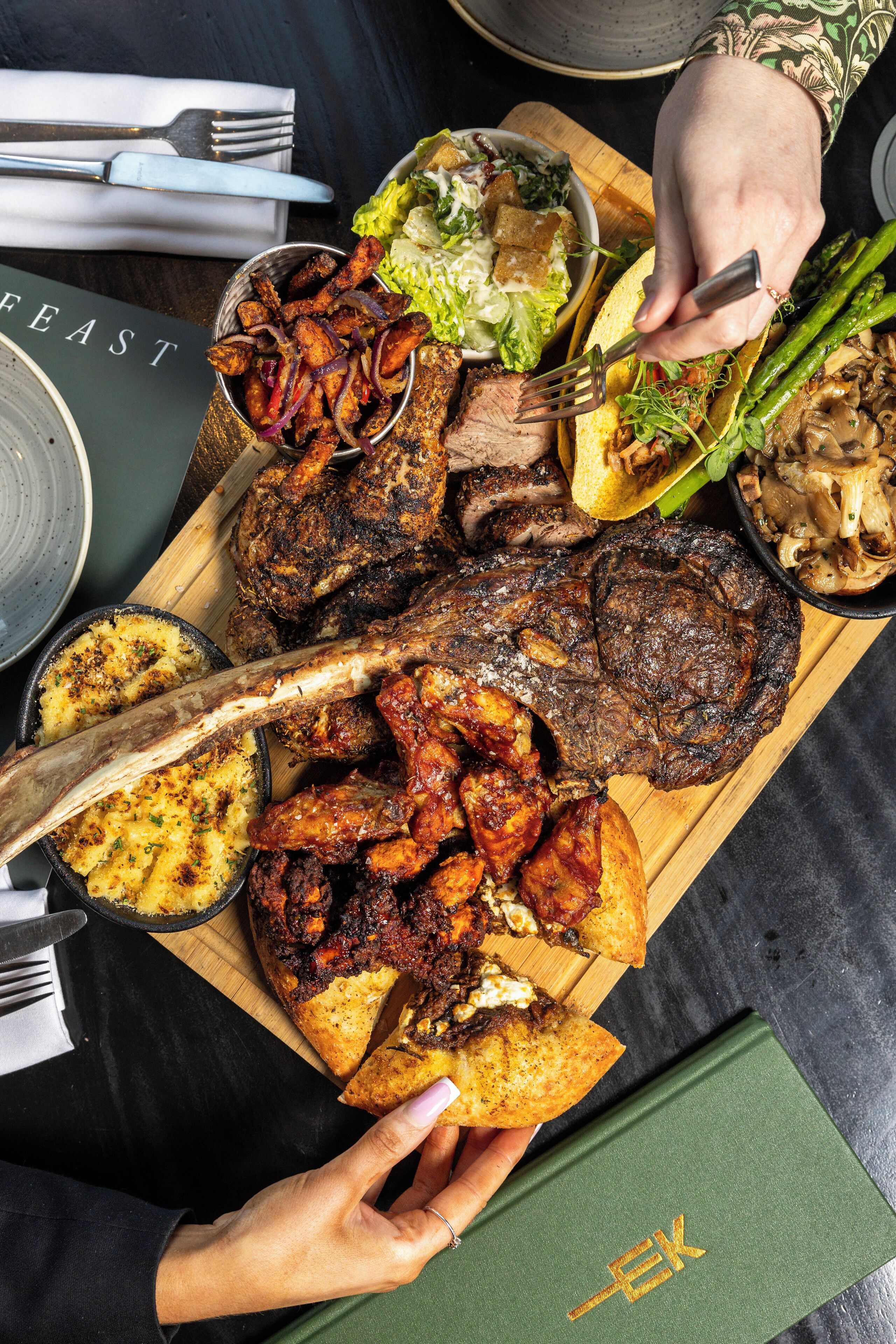 Une variété de viandes et de légumes grillés, présentés sur une planche en bois, et une personne tenant une fourchette.