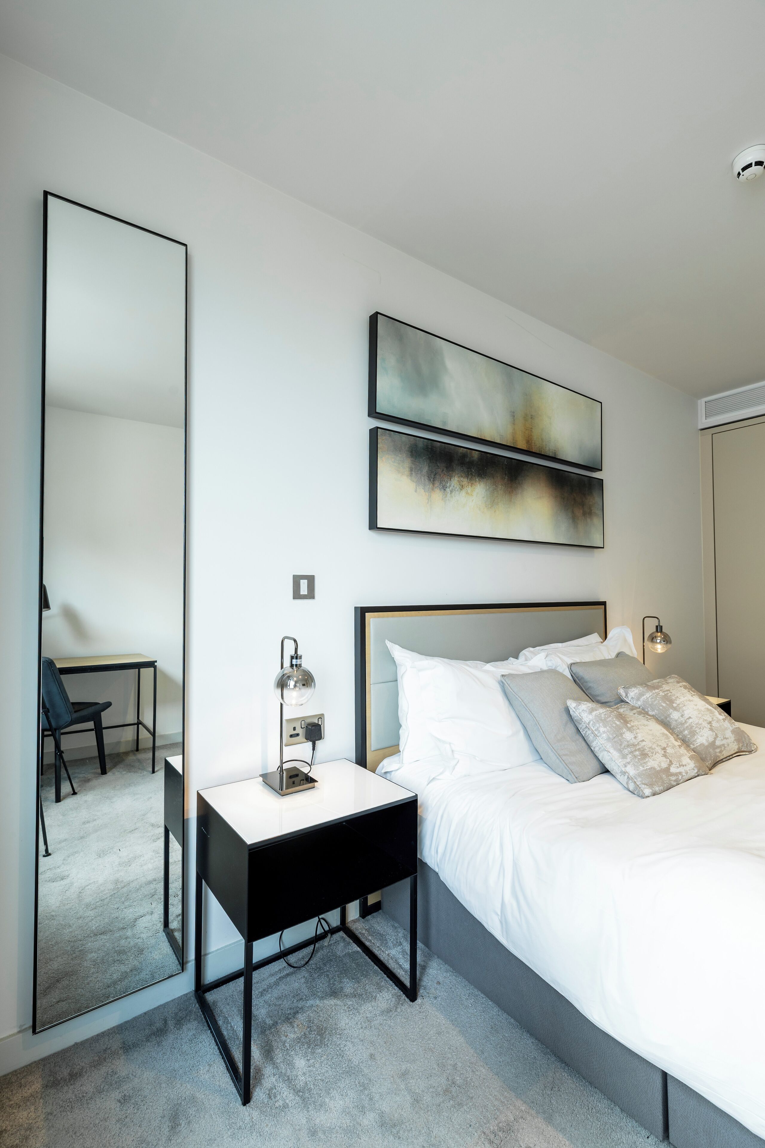A modern bedroom with a large bed, a bedside table, a desk, and a mirror.