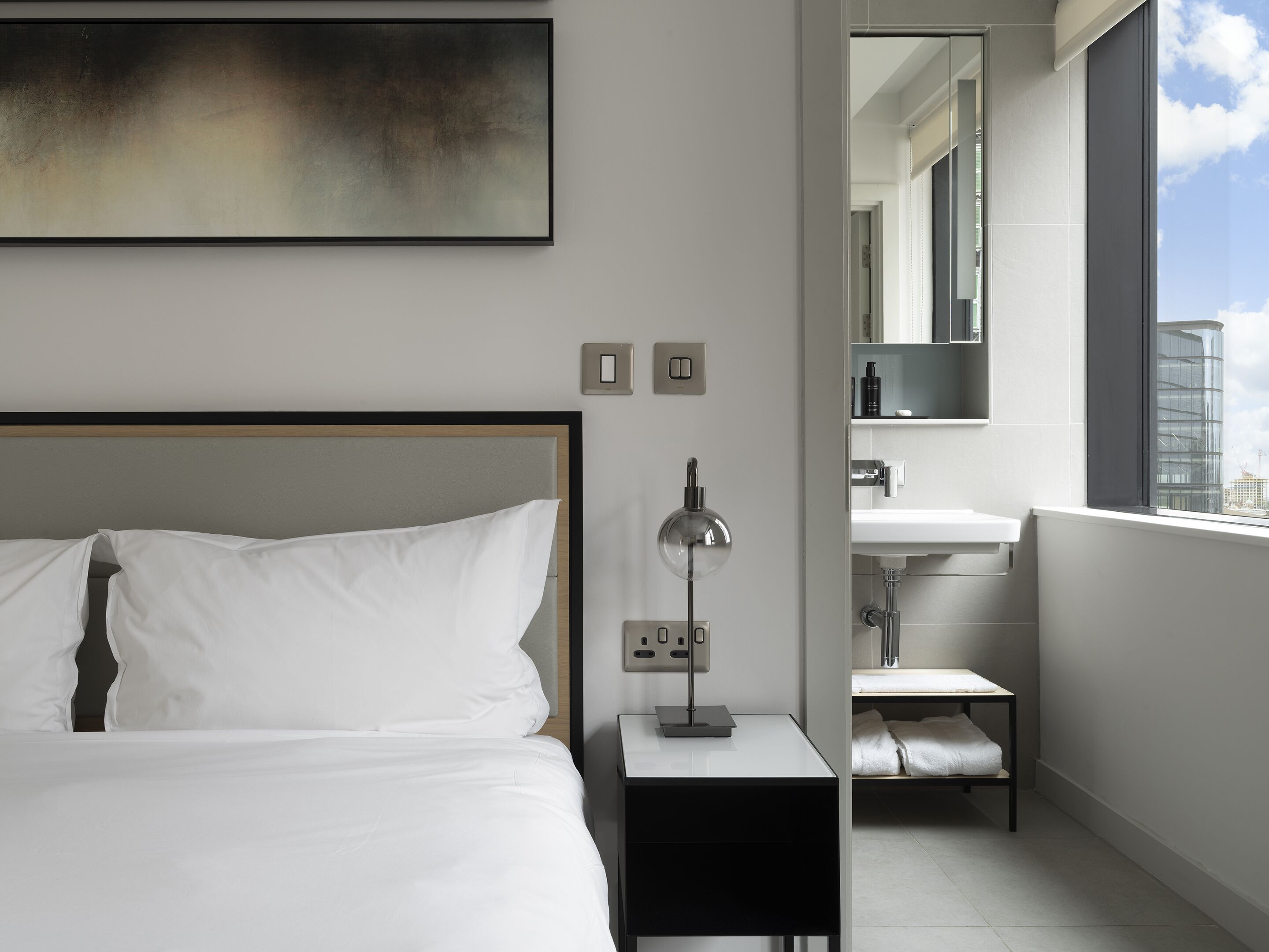 Habitación de hotel moderna con una cama grande, una mesita de noche con lámpara, una pequeña mesita de noche con teléfono, un baño con lavamanos y espejo, y vistas a la ciudad a través de un ventanal.