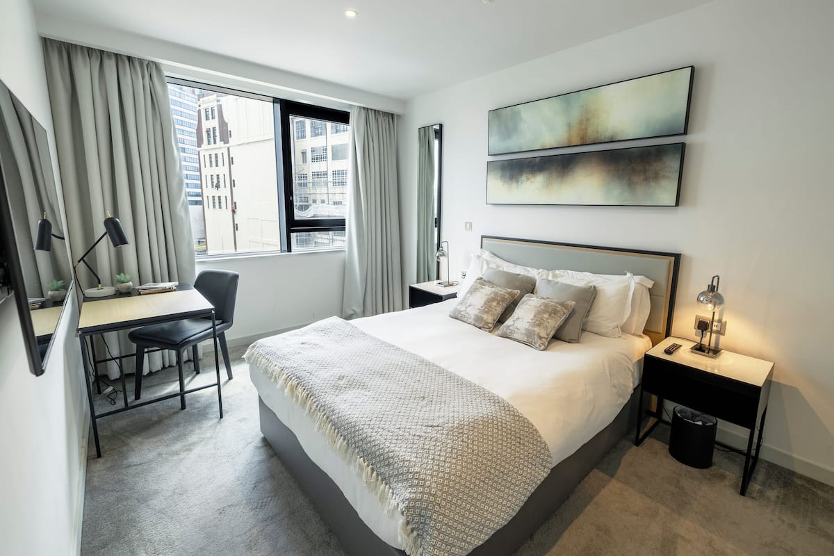 A modern hotel room with a large bed, a desk, and a chair. There are two paintings on the wall, a window with curtains, and a nightstand with a lamp.