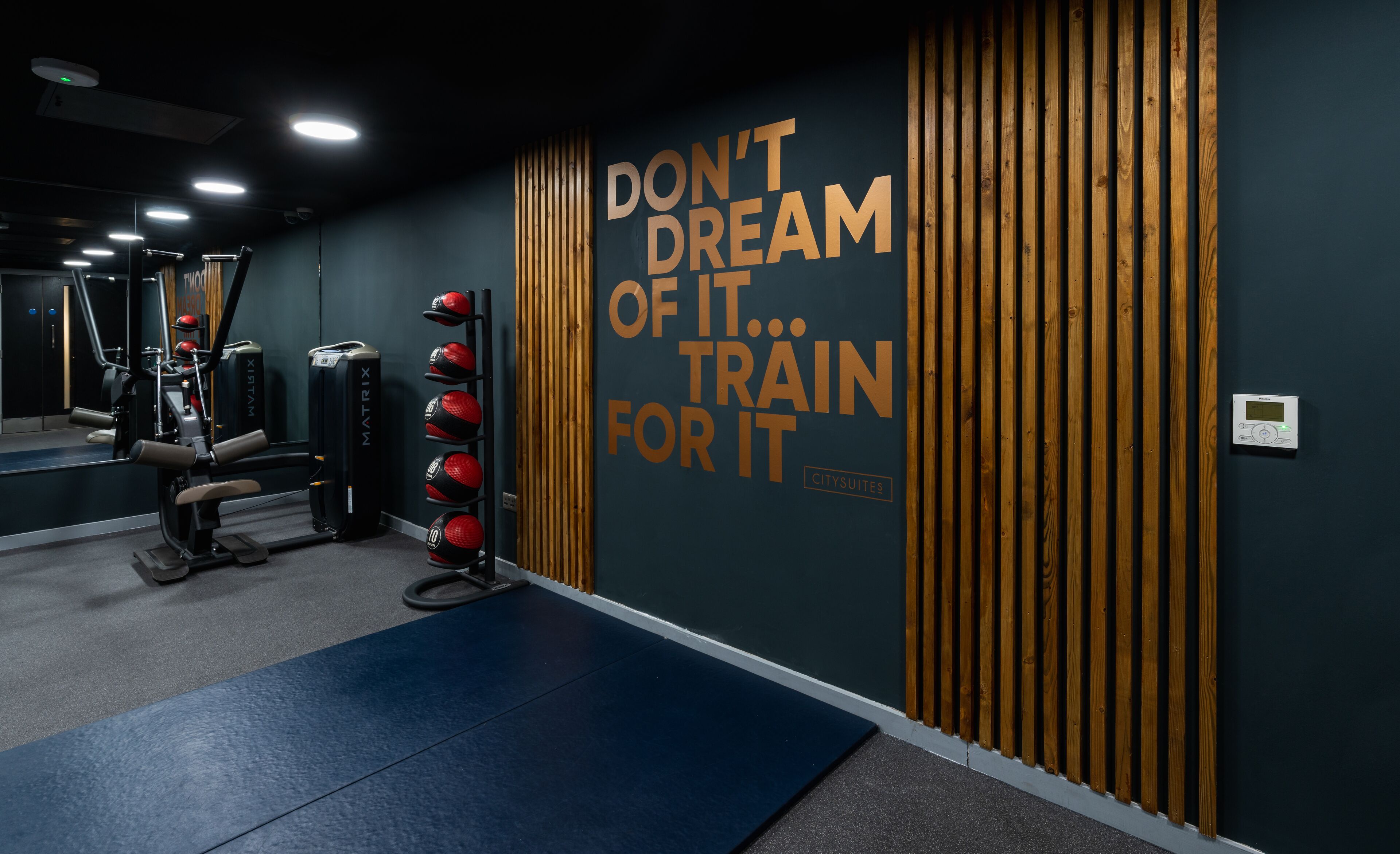 Un gimnasio con equipo de ejercicio, texto motivacional en la pared y paneles de madera.