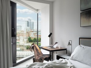 Quarto de hotel com uma grande janela, uma escrivaninha com um laptop, uma cadeira, uma mesa de cabeceira com abajur e uma cama.