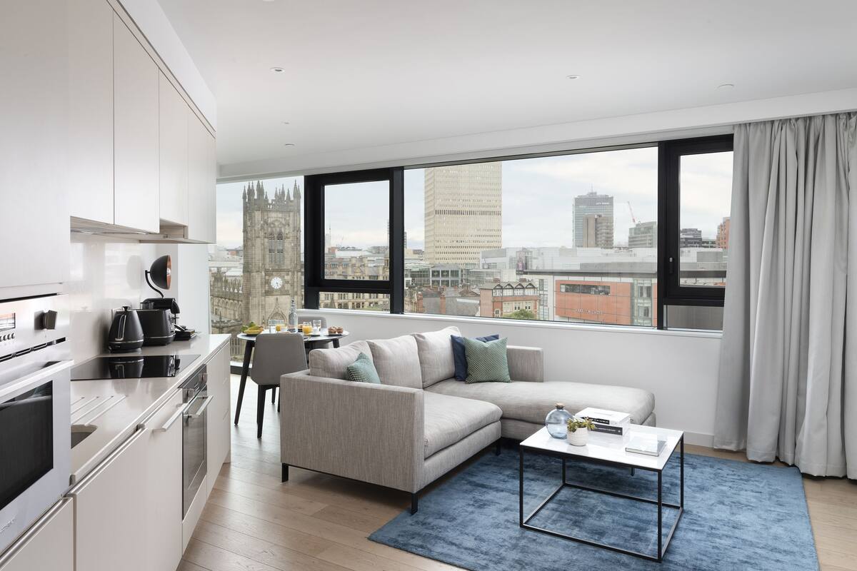 A modern apartment with a kitchen, dining area, and living room. It features a large window with a city view, a sofa, and a coffee table.