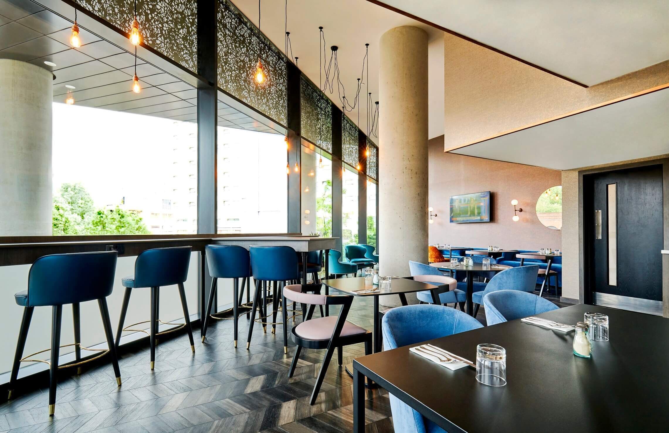 A modern restaurant with bar seating, large windows, and a view of greenery.