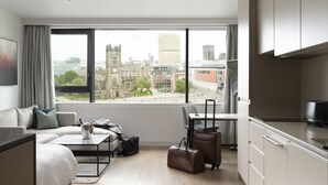 A modern hotel room with a large window, a bed, a sofa, a dining table, and a kitchenette. - CitySuites Aparthotel (Salford)