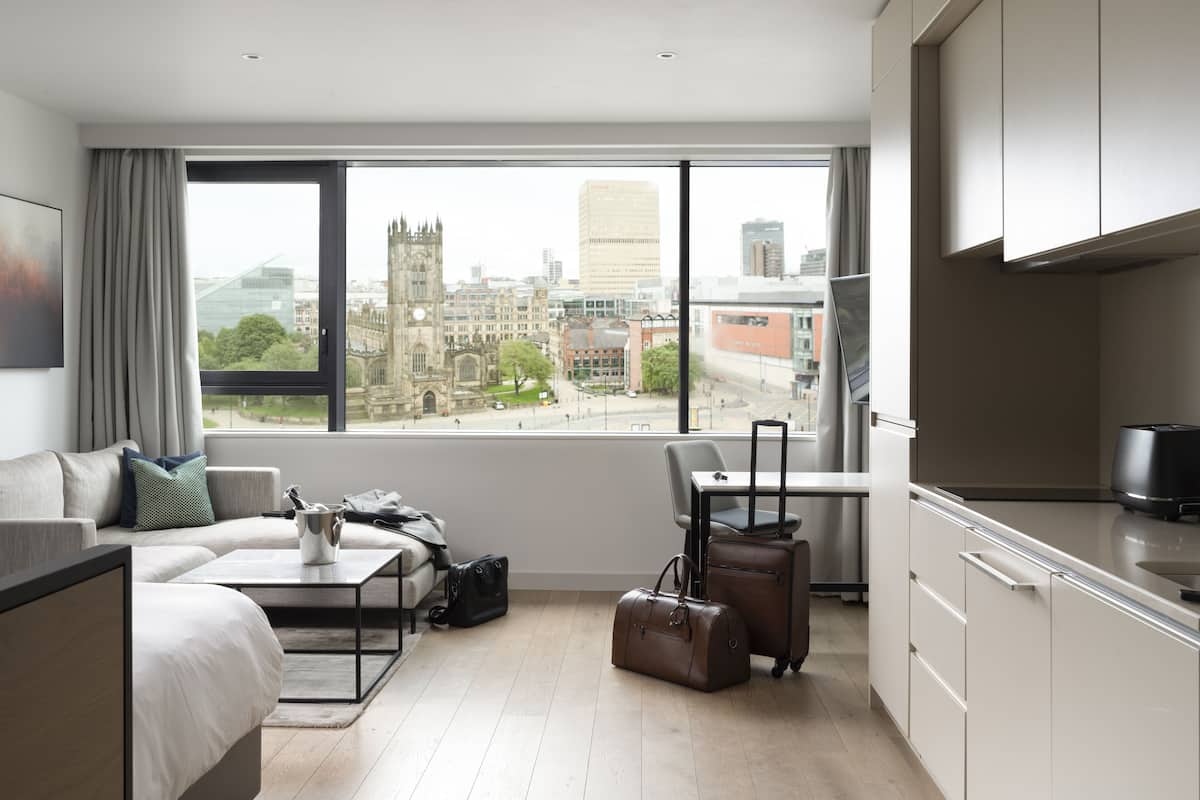 A modern hotel room with a large window, a bed, a sofa, a dining table, and a kitchenette.