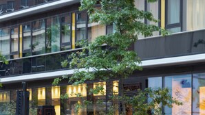 A modern hotel with a glass facade, outdoor seating, and a visible entrance. - CitySuites Aparthotel (Salford)