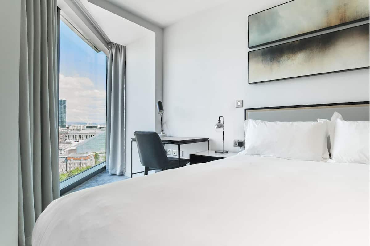 A hotel room with a large bed, a desk, a chair, and a view of the cityscape through a large window.