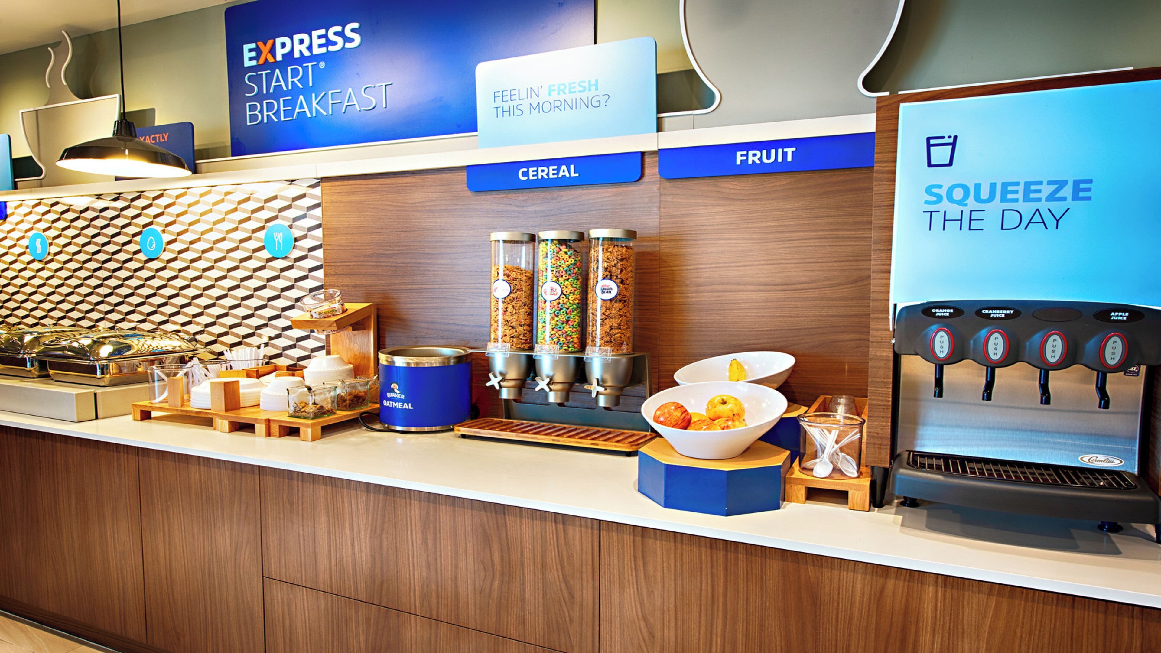 Espaço de café da manhã de hotel com sinal indicando 'EXPRESS START BREAKFAST', dispensador de cereais e frutas, dispensador de sucos e uma tigela de frutas.