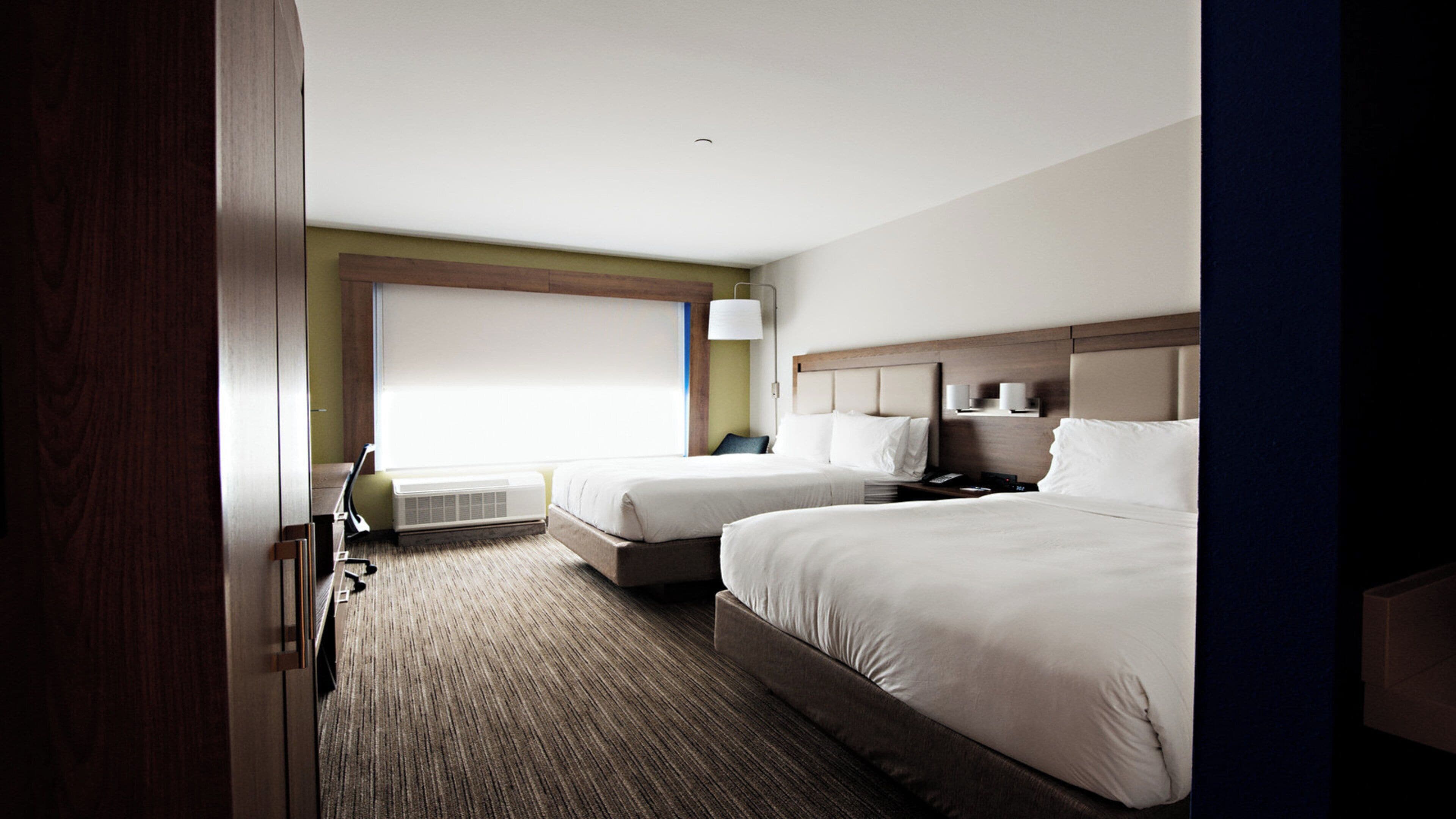 A hotel room with two beds, a large window, a wall-mounted air conditioner, and a bedside table with a lamp.