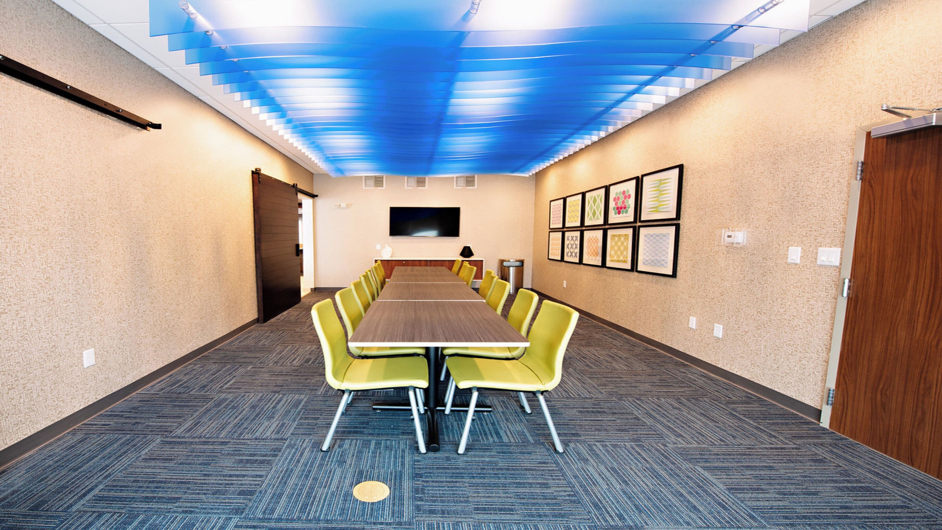A conference room with a long table, yellow chairs, a blue ceiling, and framed pictures on the wall.