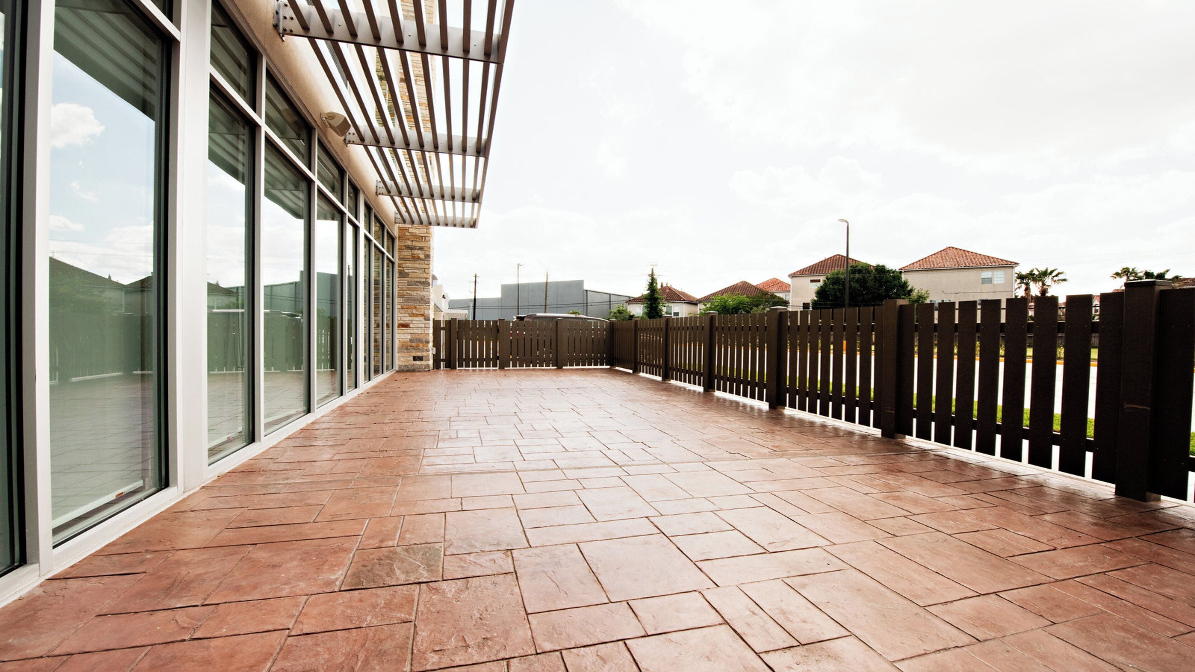 A covered outdoor area with a tiled floor, a wooden fence, and a building with large windows in the background.