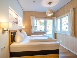 A modern bedroom with a wooden headboard, a large bed with white linens, a hanging light fixture, and a view of the outside through large windows. - Premium chalet with whirlpool (Murau)