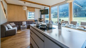 A modern kitchen with a large island, wooden beams, and large windows offering a view of the outdoors. - Premium chalet with whirlpool (Murau)