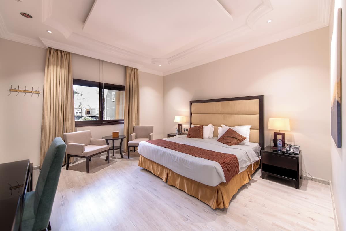 A hotel room with a large bed, two bedside tables with lamps, a desk, a chair, and a window with curtains.