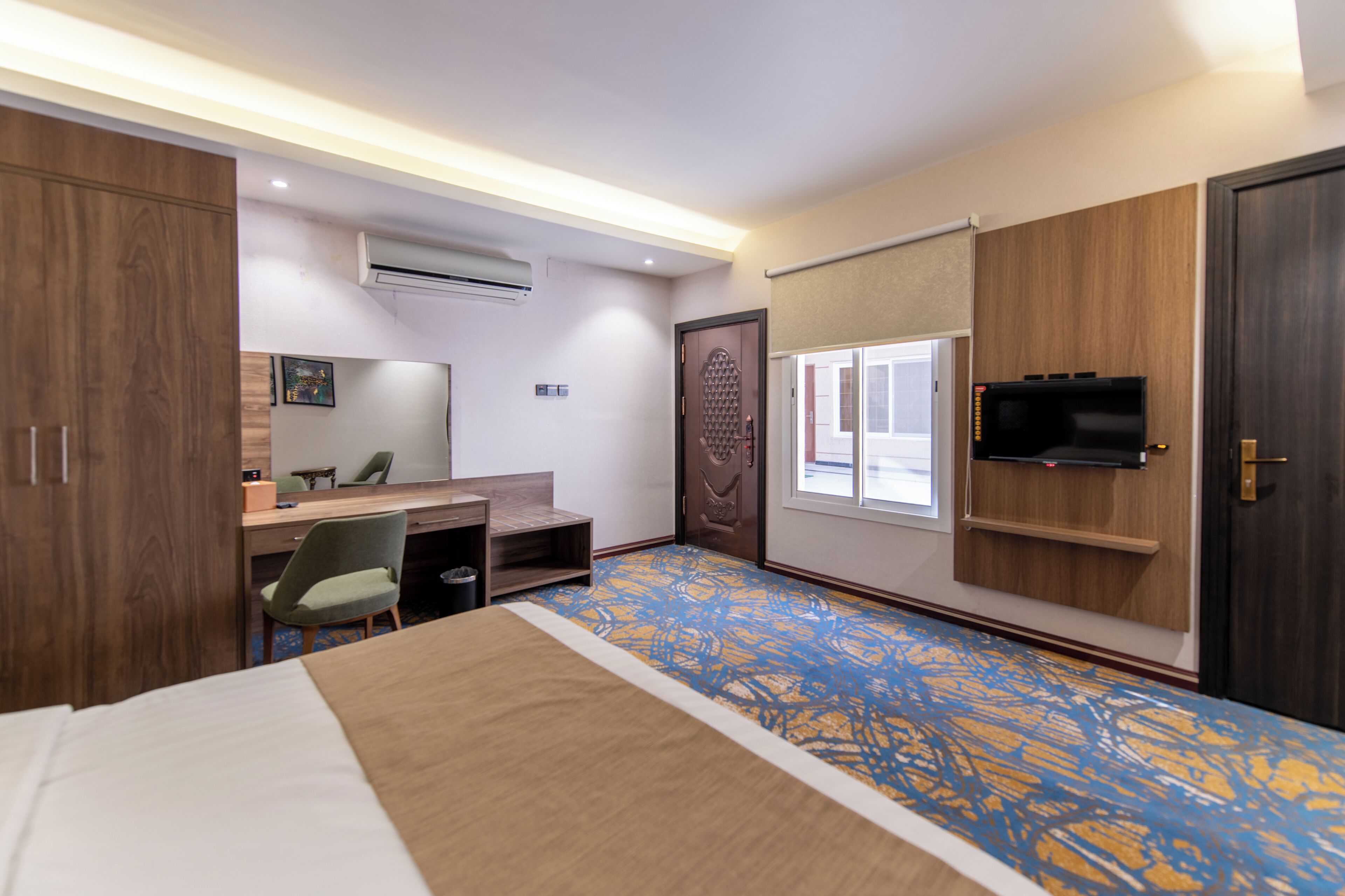 A hotel room with a bed, desk, chair, TV, wardrobe, and an air conditioning unit.