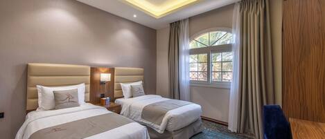 A hotel room with two beds, a desk, a chair, a window with curtains, and a ceiling light.