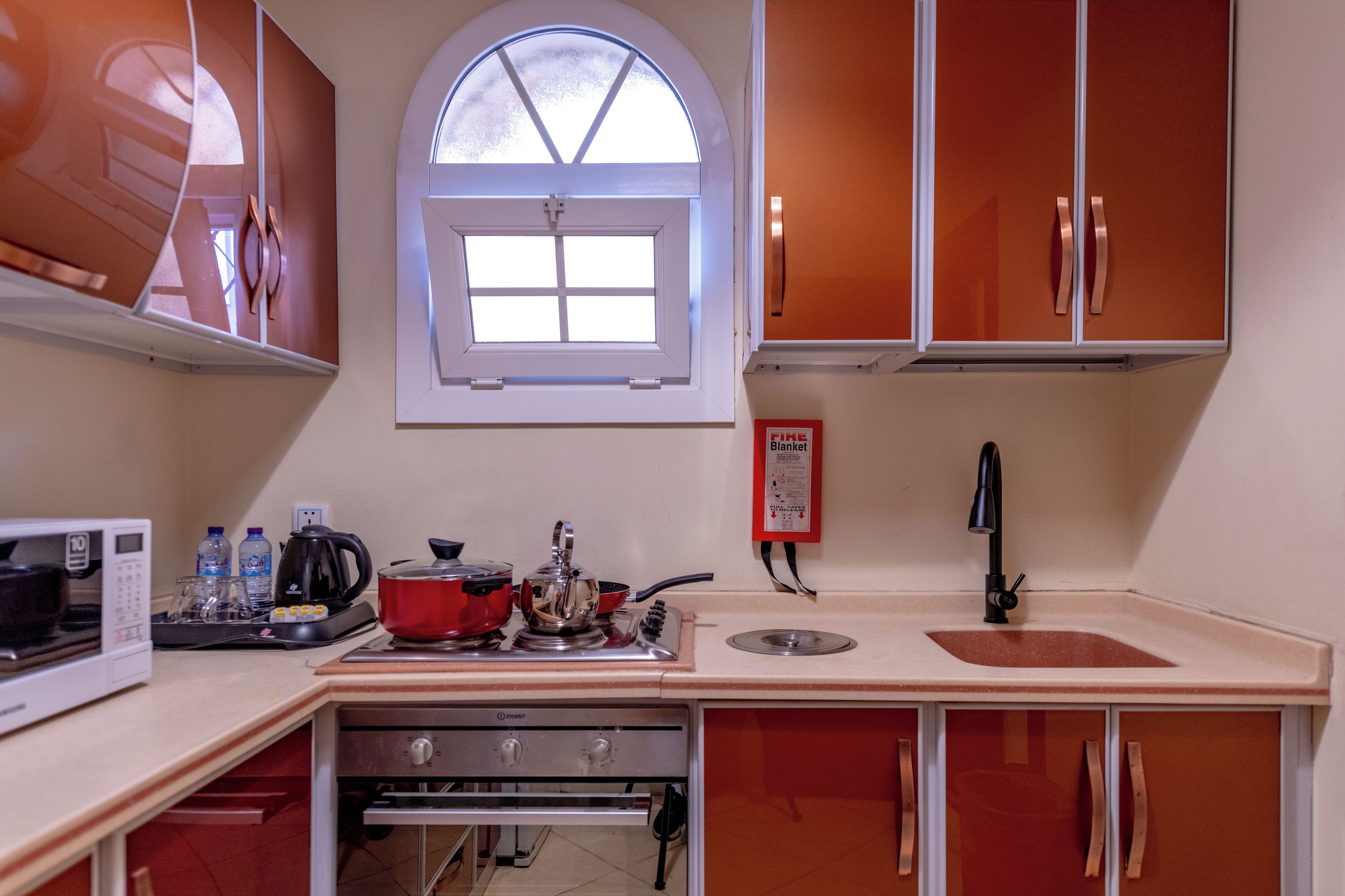 A compact kitchen with a microwave, kettle, and stove, a small sink, and a window above the sink.